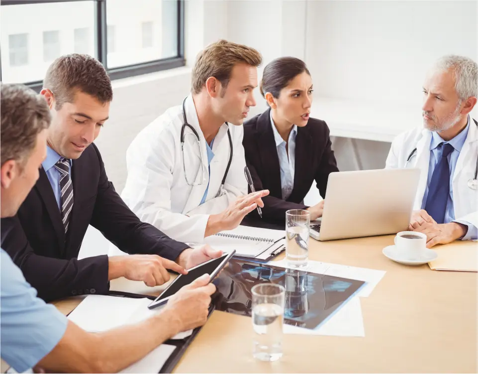 h2 (1) A group of medical professionals and businesspeople having a meeting around a table with laptops, tablets, and medical images.