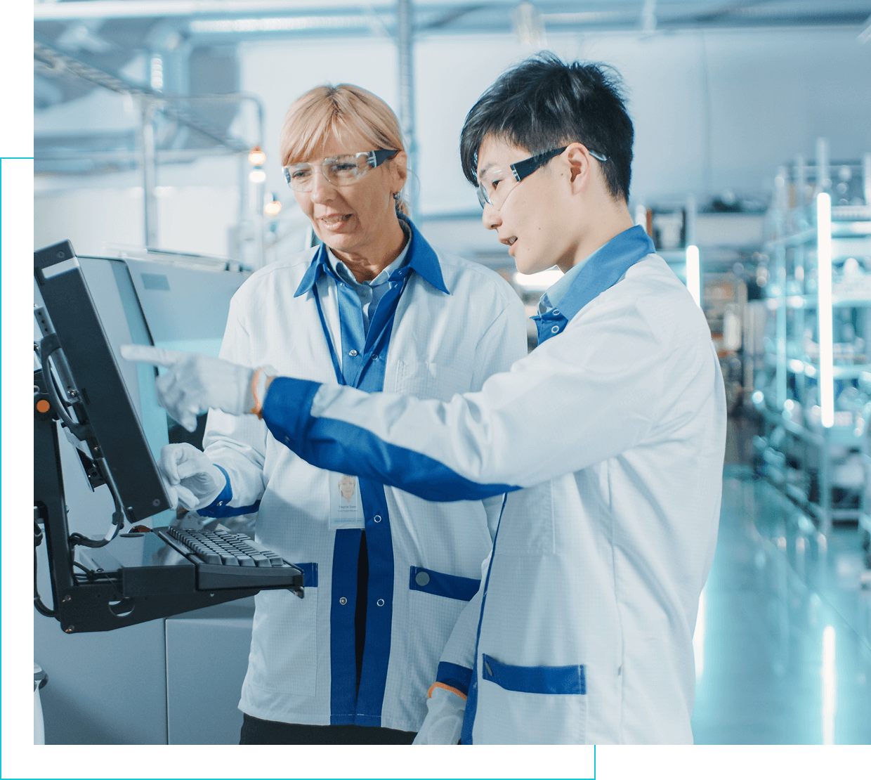 Group 1321317424 Two people in lab coats and safety glasses are looking at a monitor in an industrial setting. One points at the screen while the other observes attentively.