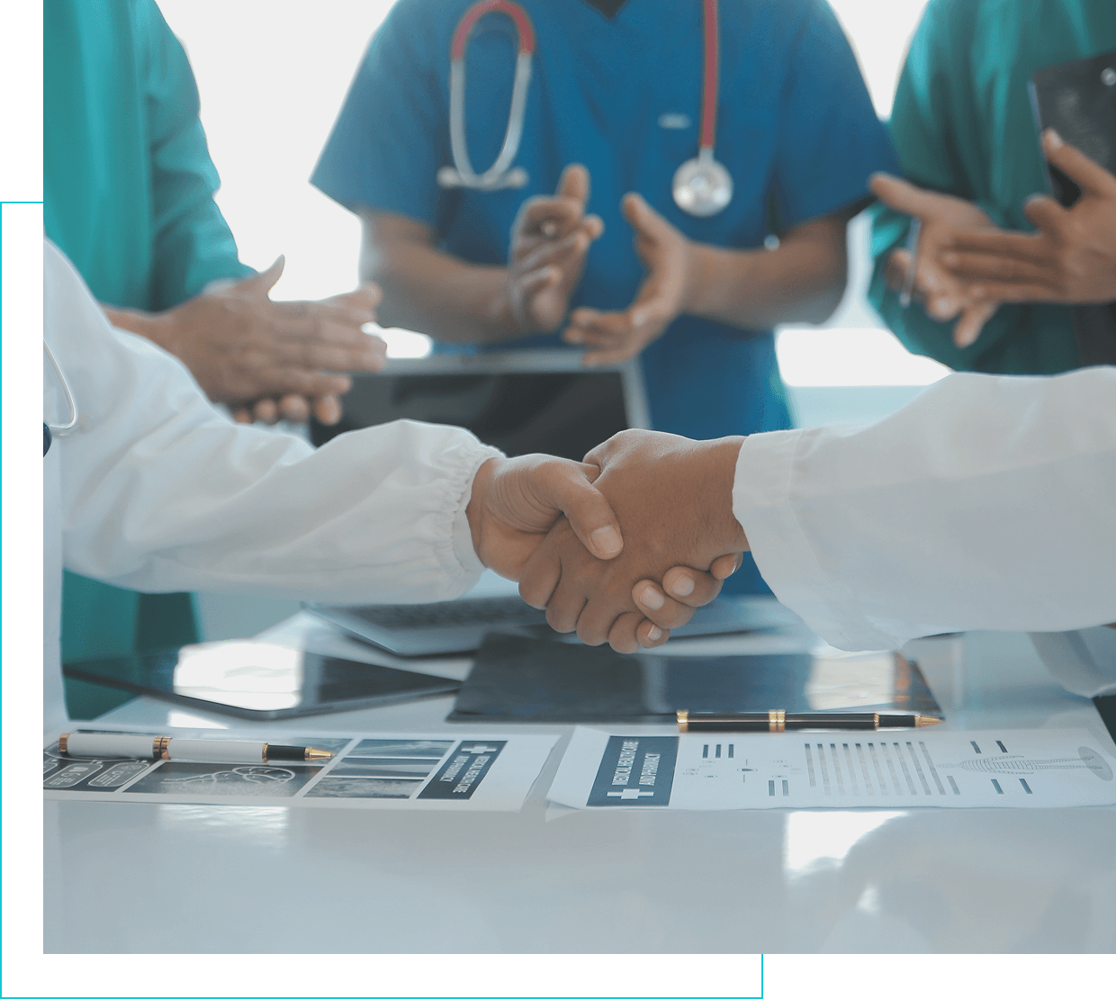 Group 1321317430 Two people in lab coats shaking hands over a table with medical documents, with other individuals in scrubs and medical attire in the background.